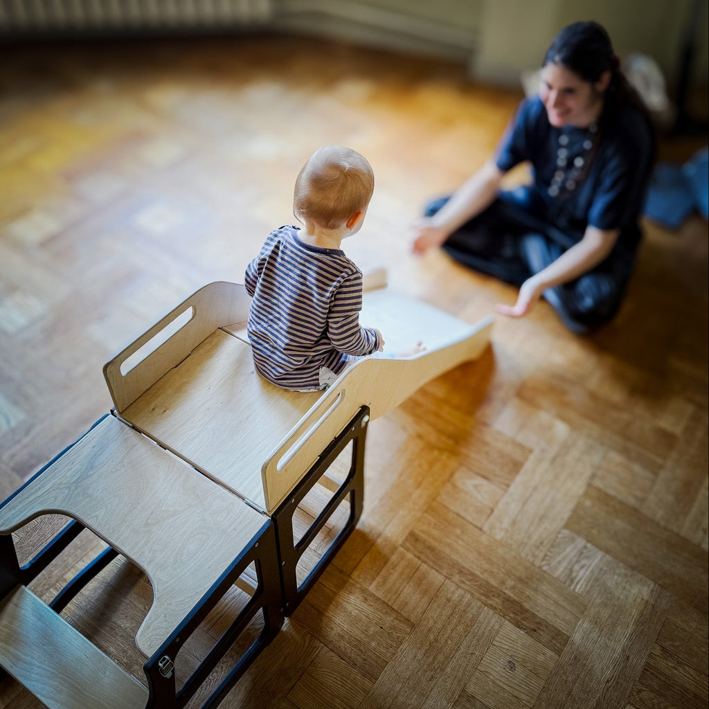 Child sitting on a wooden slide with an adult nearby in a room with wooden flooring.