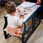 Child sitting at a small desk with books and a teddy bear, supervised by an adult.